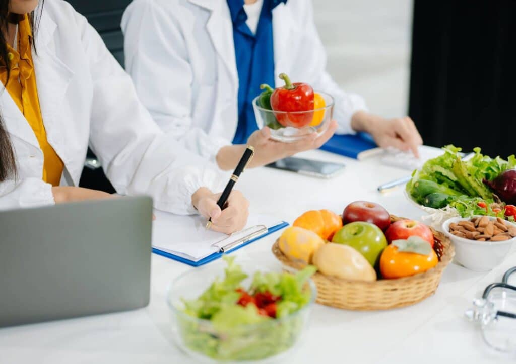Nutritionist vs Clinical Nutritionist: What's the Difference? 8 Two doctors collaborate on a laptop at a table, surrounded by a bowl of fresh fruit and vegetables.
