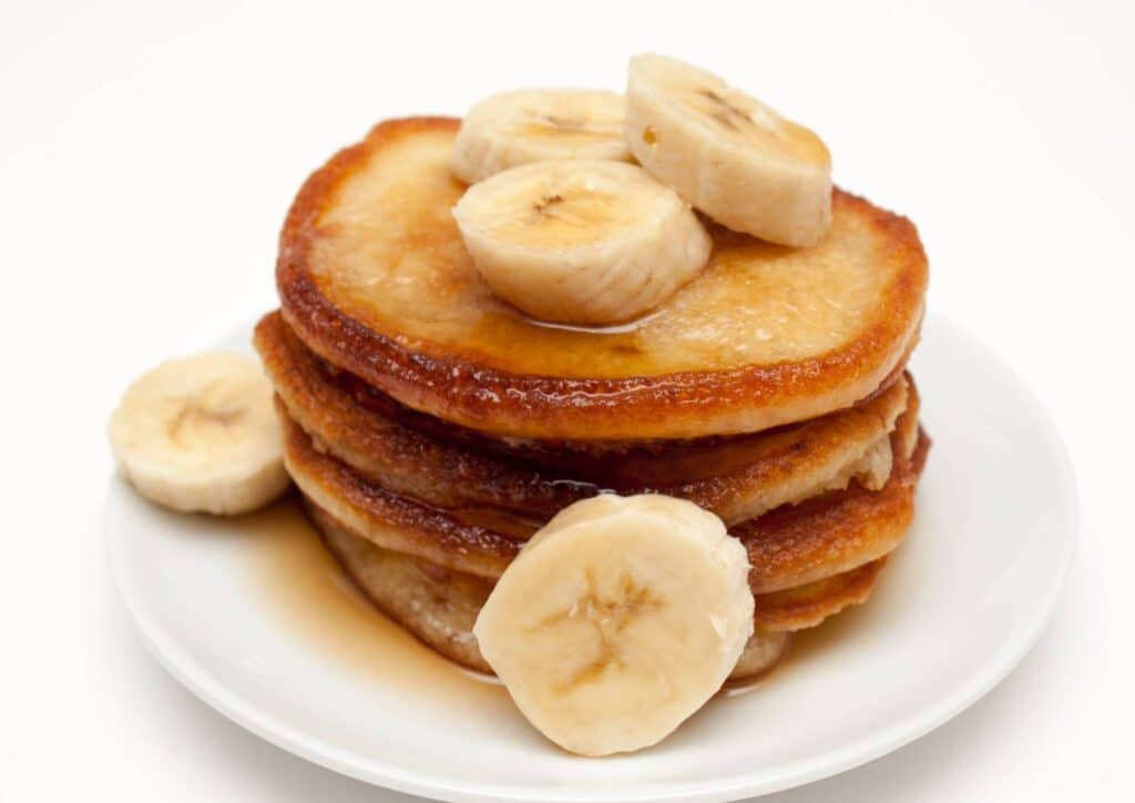 Protein Pancakes Breakfast Recipe for Weight Loss 7 A stack of golden pancakes topped with sliced bananas, served on a white plate.
