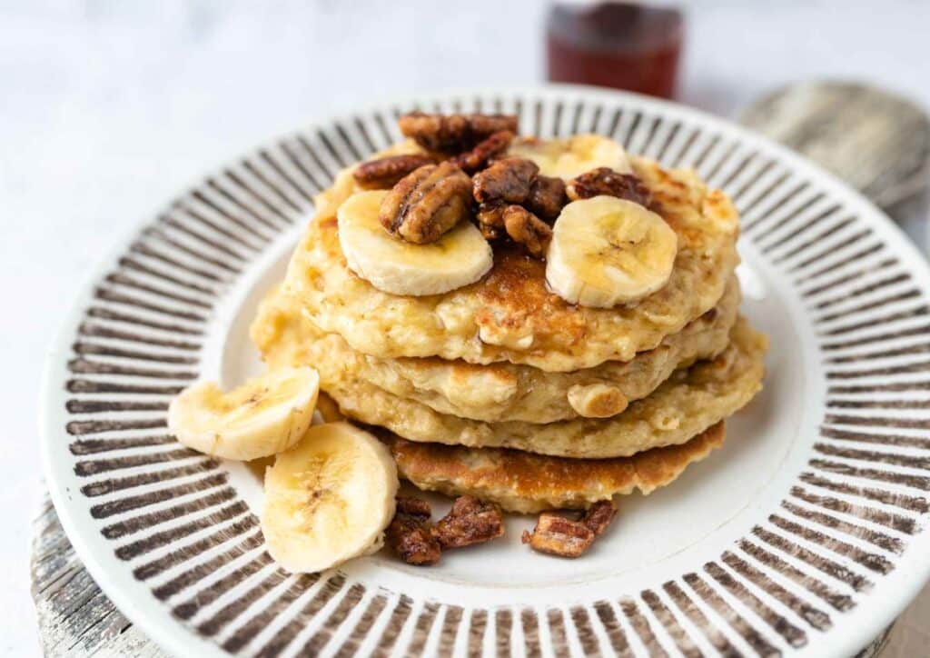 Protein Pancakes Breakfast Recipe for Weight Loss 6 A stack of pancakes topped with sliced bananas and chopped pecans, served on a white plate.