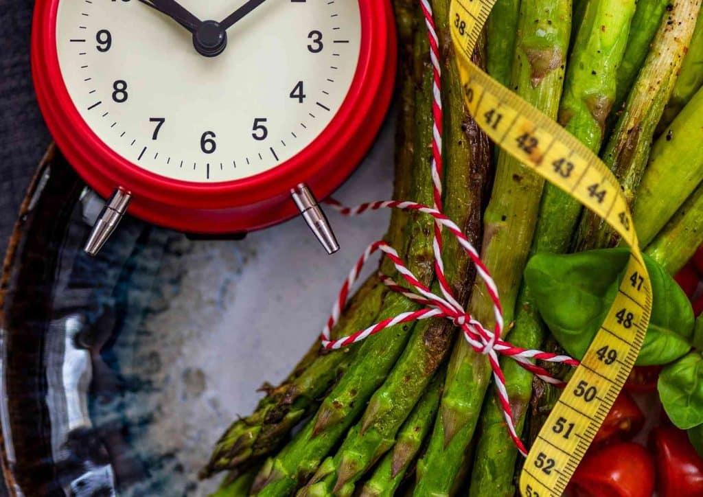 What Is Sustainable Weight Loss? 4 Fresh asparagus and tomatoes beside a red alarm clock on a wooden surface, suggesting a meal preparation theme.