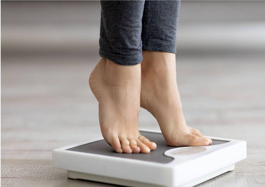 What Is Sustainable Weight Loss? 14 A woman stands on a scale, looking down at the display with her feet positioned firmly on the scale's surface.
