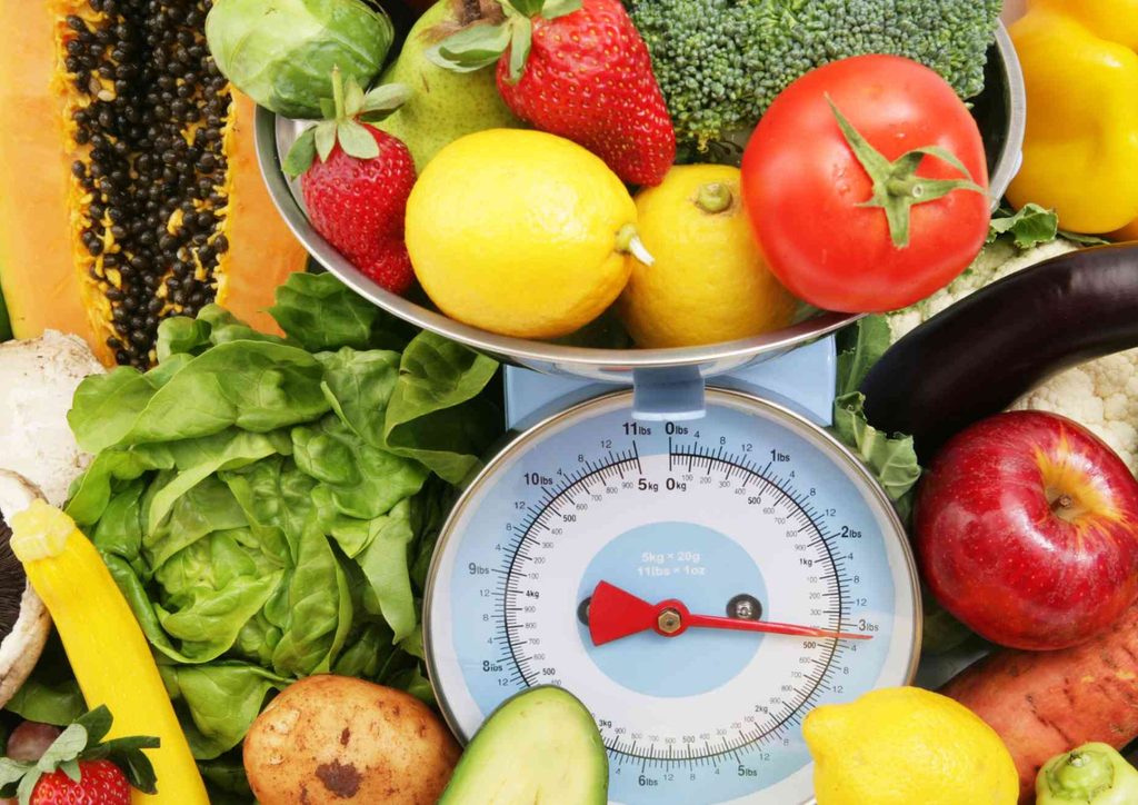Not Losing Weight in a Calorie Deficit? Here's Why 26 Fresh fruits and vegetables arranged on a scale, indicating their combined weight for nutritional assessment.