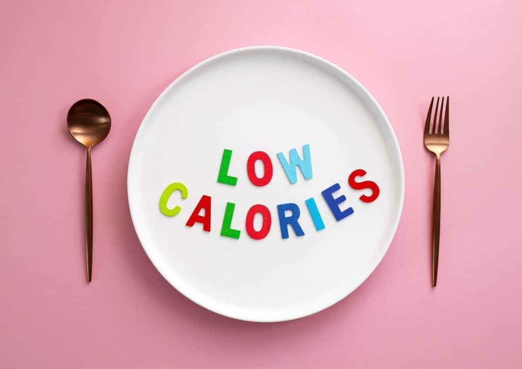 Not Losing Weight in a Calorie Deficit? Here's Why 2 The phrase "low calories" displayed on a white plate, accompanied by a fork and spoon, set against a pink background.