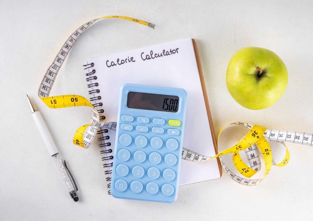 Not Losing Weight in a Calorie Deficit? Here's Why 5 A calculator, measuring tape, apple, and notebook arranged together on a surface, indicating a study or measurement context.