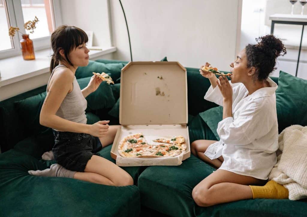 Not Losing Weight in a Calorie Deficit? Here's Why 13 Two women sitting on a couch, enjoying pizza from a box, smiling and sharing a casual moment together.