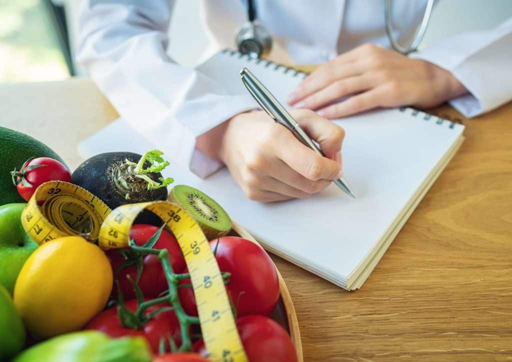 How to Choose the Best Weight Loss Nutritionist in Sydney 26 A doctor writes notes on a notepad beside a bowl filled with fresh fruits and vegetables, promoting healthy eating.