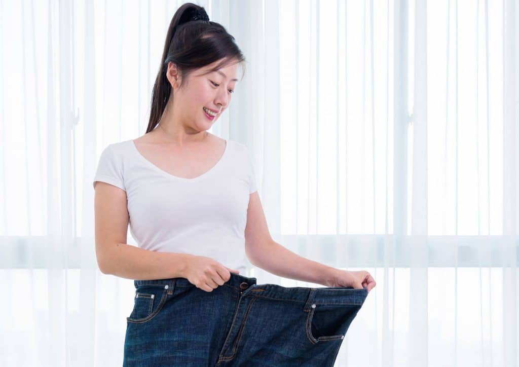 How to Lose Weight Without Exercise 7 Asian woman in a white shirt and blue jeans, holding up her jeans with a smile, showcasing her outfit.