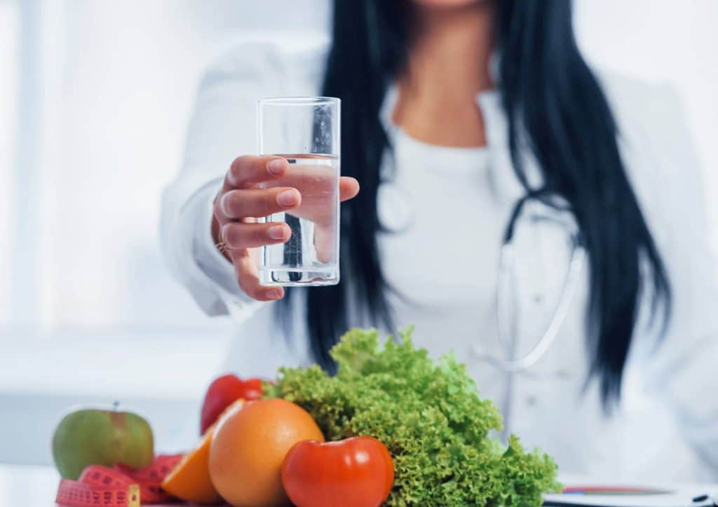 How to Choose the Best Weight Loss Nutritionist in Sydney 5 A woman holds a glass of water surrounded by fresh fruits and vegetables, promoting healthy hydration and nutrition.