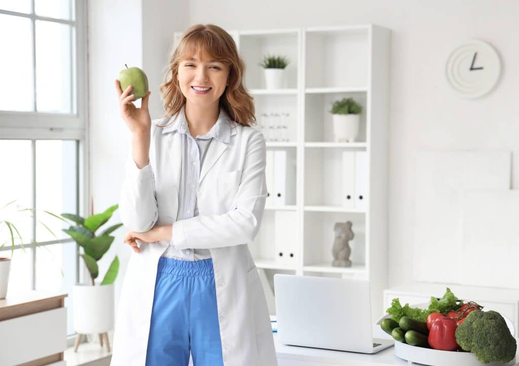 How to Choose the Best Weight Loss Nutritionist in Sydney 13 A cheerful woman in a lab coat presents a fresh apple, representing her focus on health and wellness.