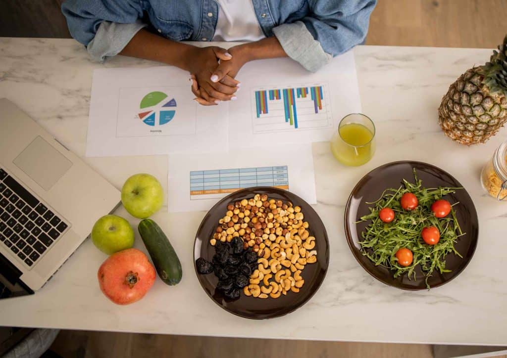 How to Choose the Best Weight Loss Nutritionist in Sydney 3 A woman sits at a table with a laptop, flanked by two plates of food, engaged in a meal while working.
