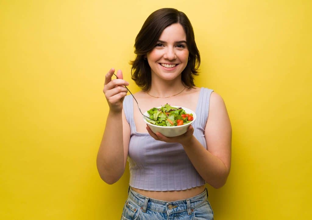 How to Lose Weight Without Exercise 9 A woman holds a bowl of fresh salad against a vibrant yellow background, showcasing healthy eating.