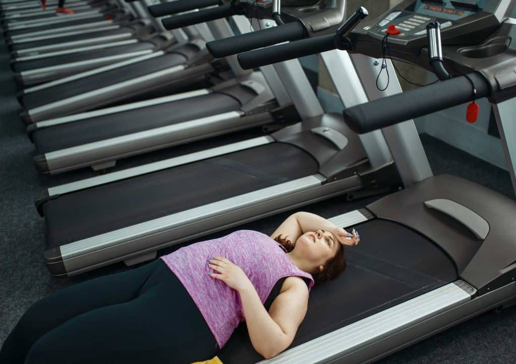 How to Lose Weight Without Exercise 1 A woman lies on a treadmill in a gym, appearing fatigued and resting after a workout session.