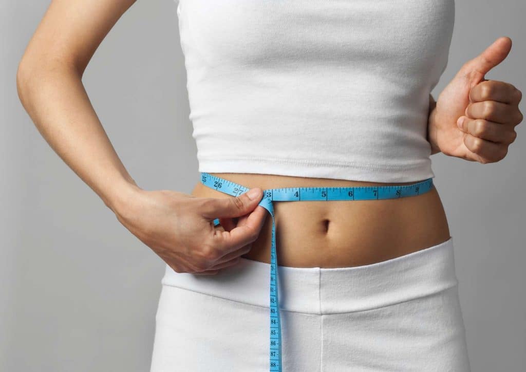 How to Lose Weight Without Exercise 17 A woman measuring her waist with a tape measure, focusing on her midsection in a well-lit room.