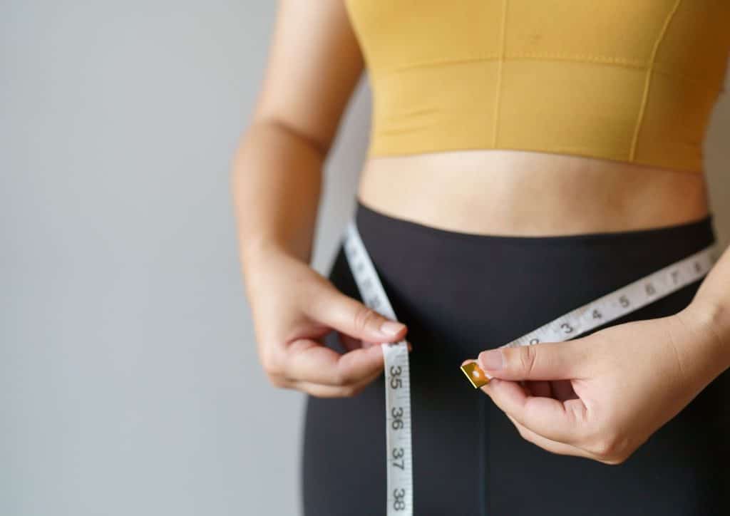 How to Lose Weight Without Exercise 21 A woman measures her waist with a tape measure, focusing on her midsection in a well-lit room.