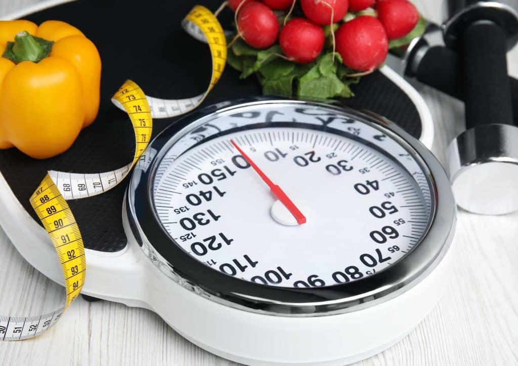 How to Lose Weight Without Exercise 12 A scale and measuring tape placed beside a variety of fresh vegetables on a wooden surface.