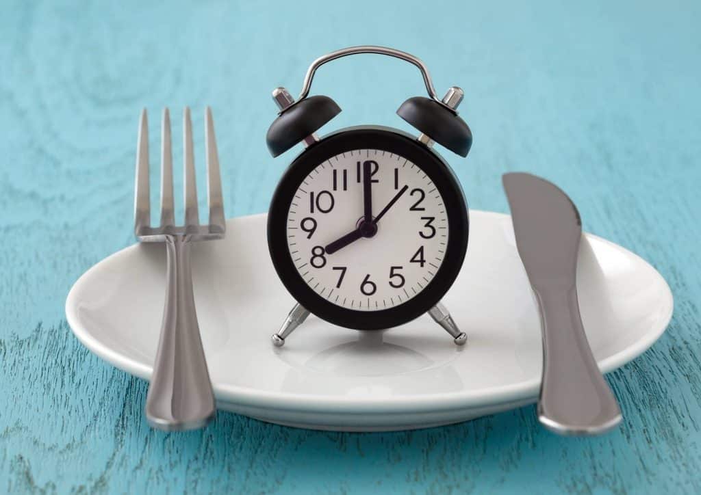 Intermittent Fasting for Weight Loss: Does It Actually Work? 19 A round clock resting on a plate, with a fork and knife positioned beside it, representing the intersection of time and meals.