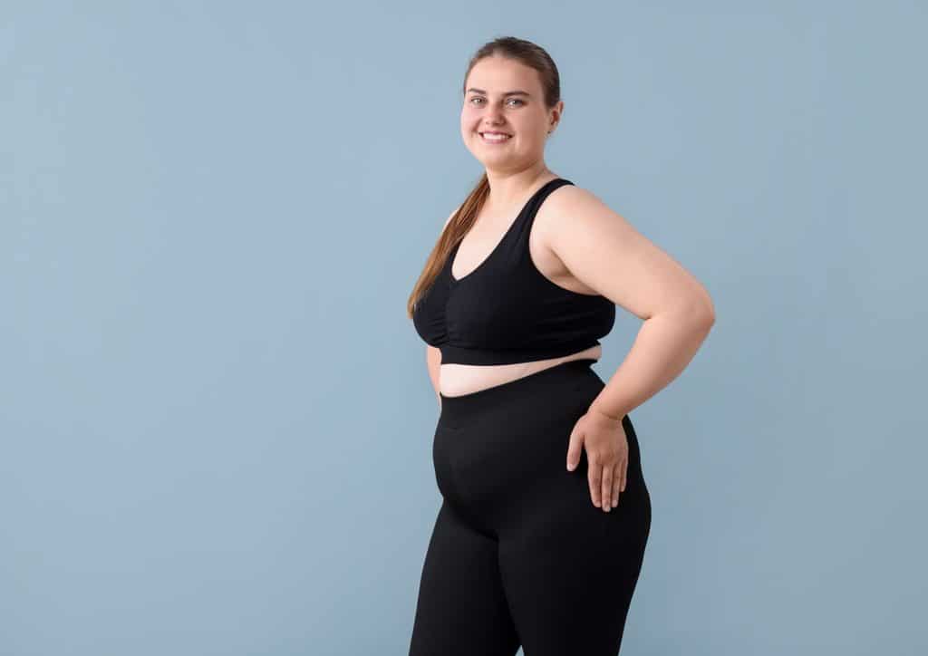 How to Lose Weight Without Exercise 26 A woman in a sports bra and shorts holds a glass of water while eating a piece of fruit, promoting a healthy lifestyle.