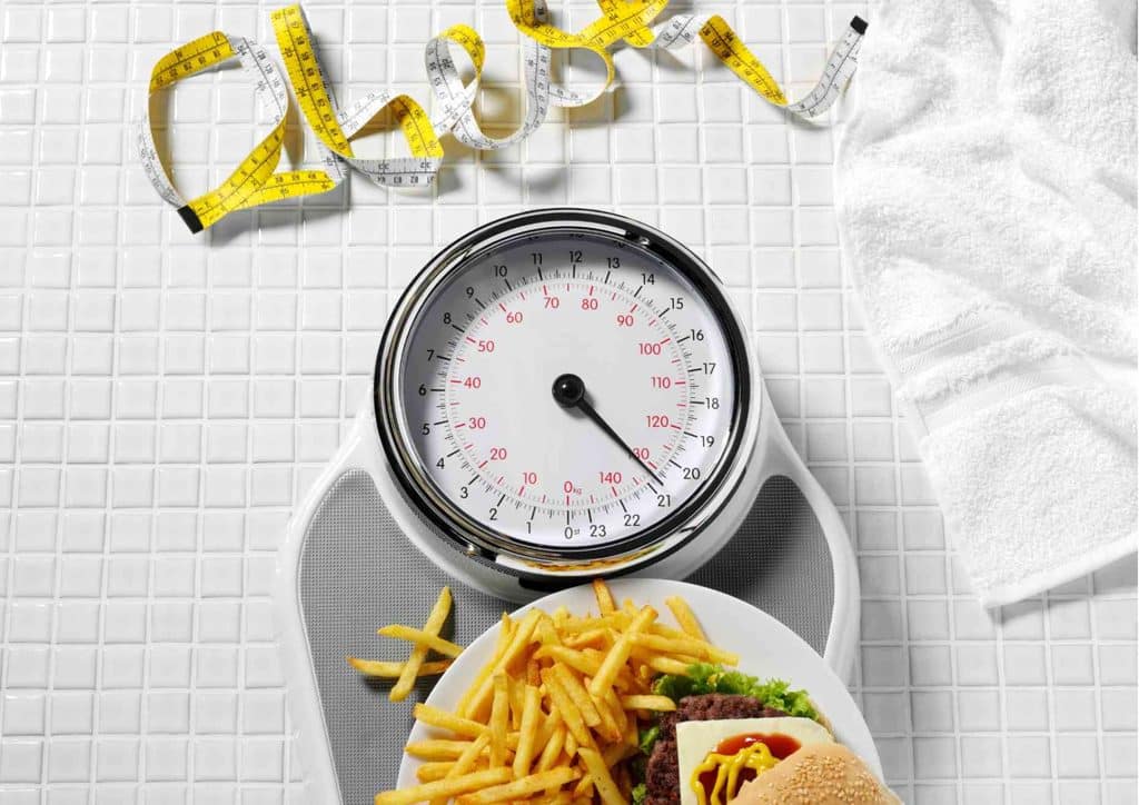 How to Lose Weight Without Exercise 22 A scale displaying a hamburger and fries, accompanied by the word 'cheat,' representing a guilty pleasure in food choices.