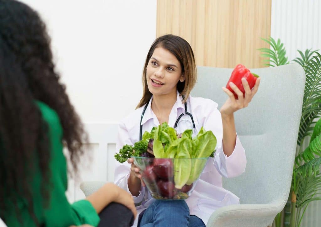 How to Choose the Best Weight Loss Nutritionist in Sydney 18 A woman sits in a chair while a doctor presents a bowl of fresh vegetables, discussing healthy eating options.