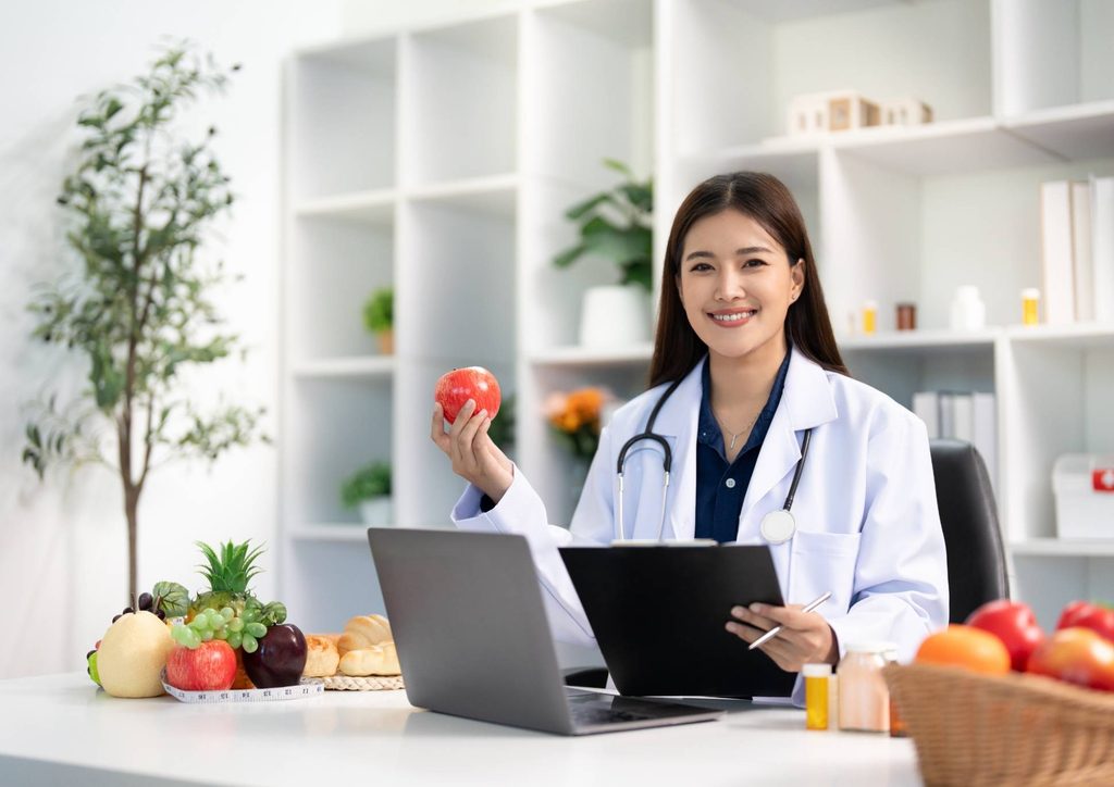 How to Choose the Best Weight Loss Nutritionist in Sydney 14 Asian female doctor in an office, holding an apple in one hand and a laptop in the other, smiling confidently.