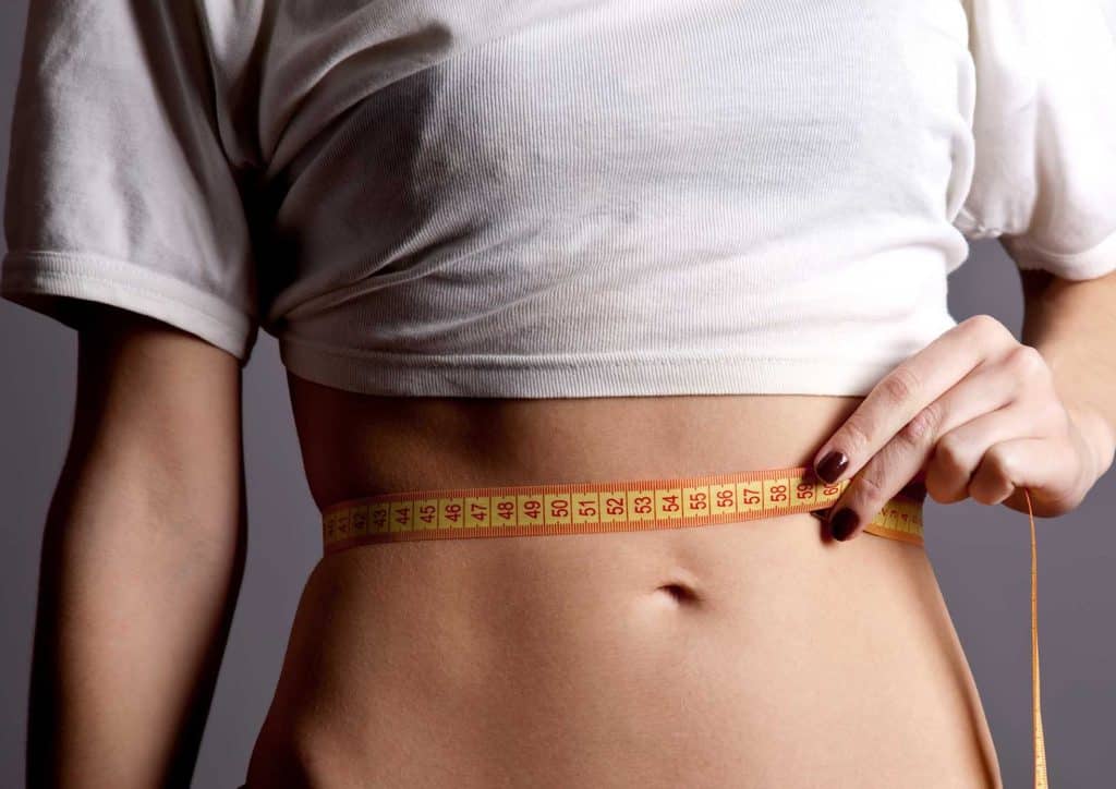 Natural Alternatives to Ozempic for Weight Loss 5 A woman measures her waist with a tape measure, focusing on her midsection in a well-lit room.