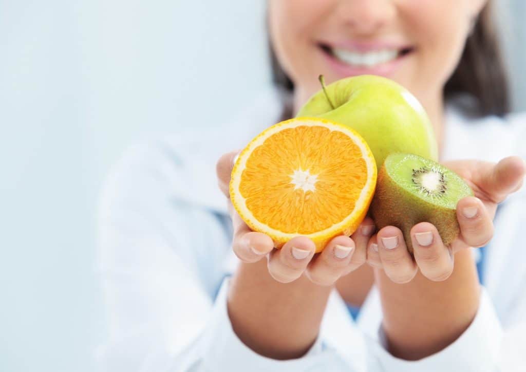 How to Choose the Best Weight Loss Nutritionist in Sydney 25 A woman in a white coat holds an orange in one hand and an apple in the other, smiling at the camera.