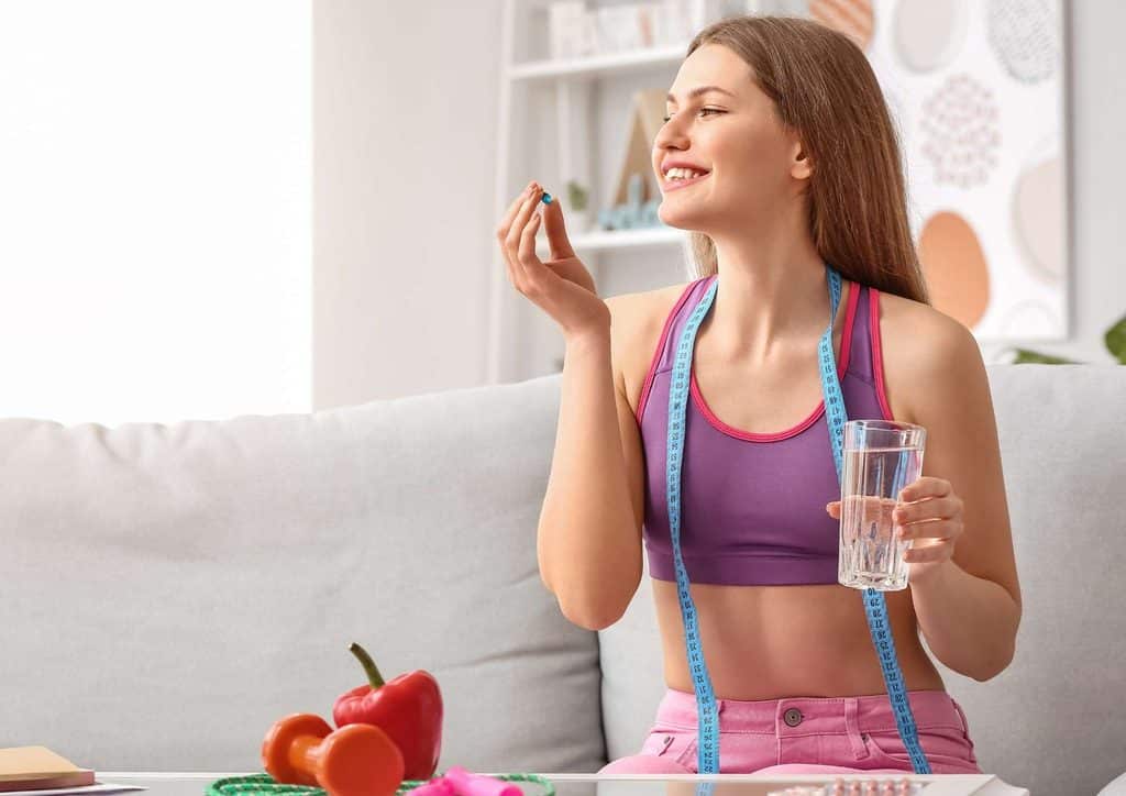 How to Lose Weight Without Exercise 28 A woman in a sports bra and shorts holds a glass of water while eating a piece of fruit, promoting a healthy lifestyle.