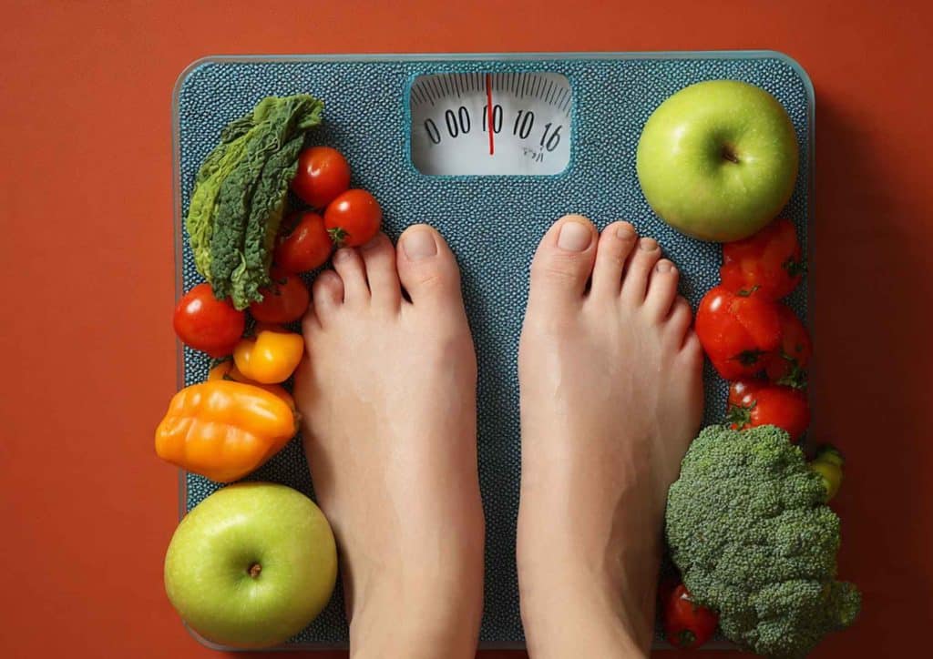 How to Lose Weight Without Exercise 25 A person's feet on a scale, surrounded by various fruits and vegetables, indicating a focus on health and nutrition.