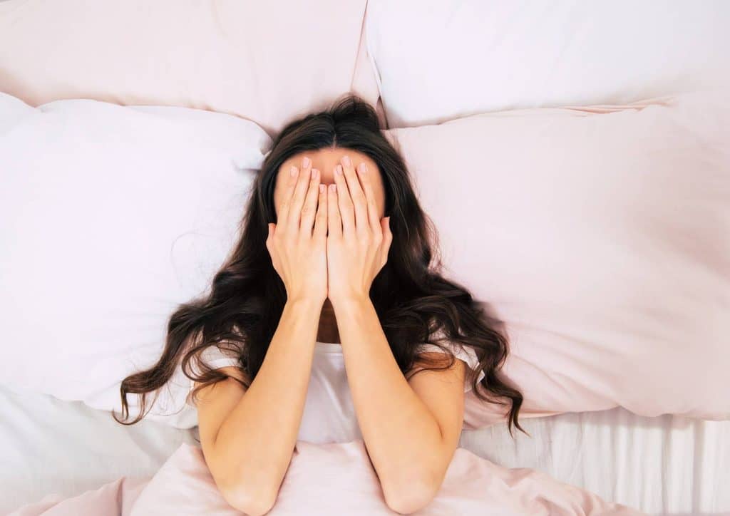 Why Am I Not Losing Weight? 8 Reasons a Nutritionist Sees Every Week 14 A woman lies in bed, covering her face with her hands, conveying a sense of distress or contemplation.