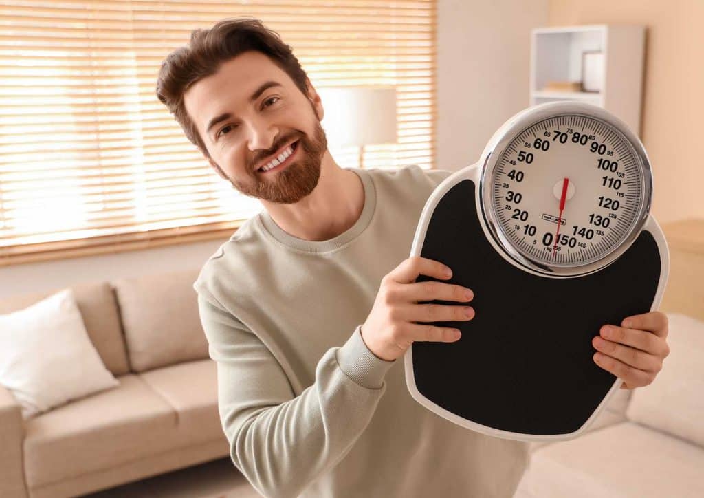 Why Am I Not Losing Weight? 8 Reasons a Nutritionist Sees Every Week 25 A man stands in his living room, holding a weight scale, looking focused and determined.