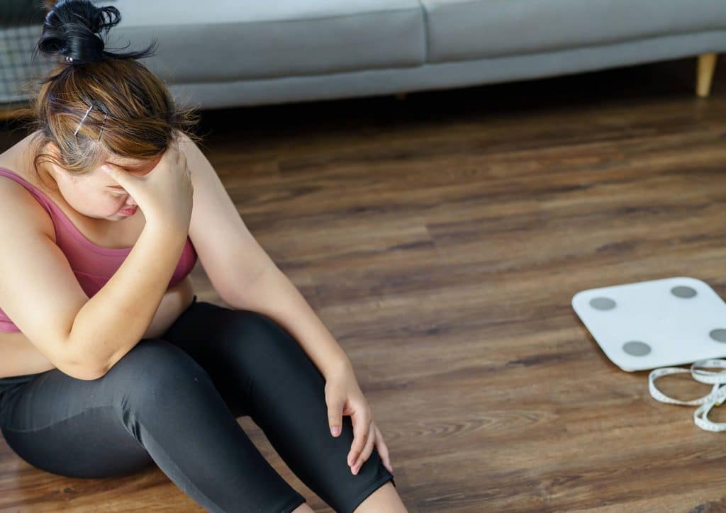 Why Am I Not Losing Weight? 8 Reasons a Nutritionist Sees Every Week 2 A woman sits on the floor beside a scale, appearing contemplative and focused on her weight measurement.