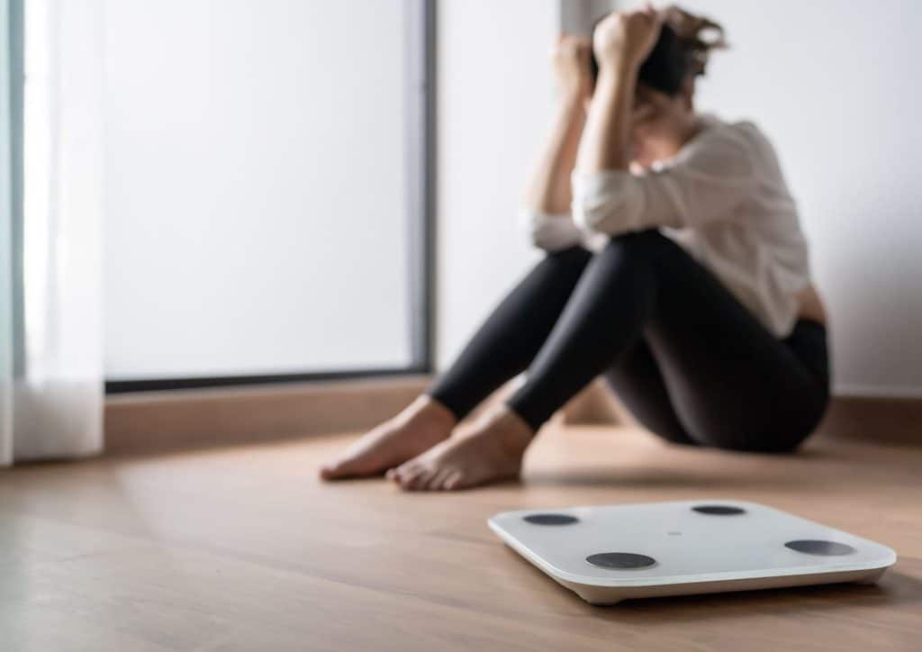 Why Am I Not Losing Weight? 8 Reasons a Nutritionist Sees Every Week 4 A woman sitting on the floor next to a scale, appearing contemplative and focused on her weight measurement.