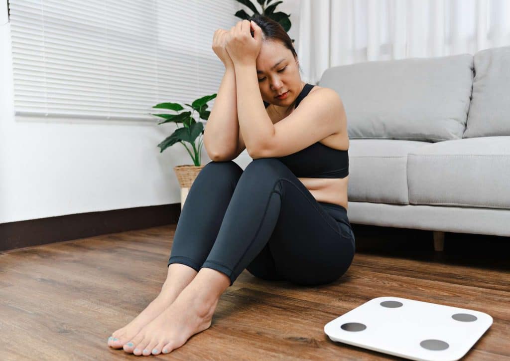 Why Am I Not Losing Weight? 8 Reasons a Nutritionist Sees Every Week 1 A woman sitting on the floor, hands on her head, appears to be in a moment of distress or contemplation.