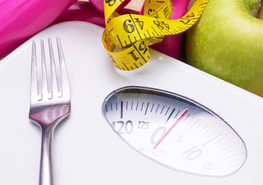 What Is a Balanced Diet for Weight Loss? 1 A fork, spoon, and an apple placed on a kitchen scale, indicating weight measurement for food preparation.