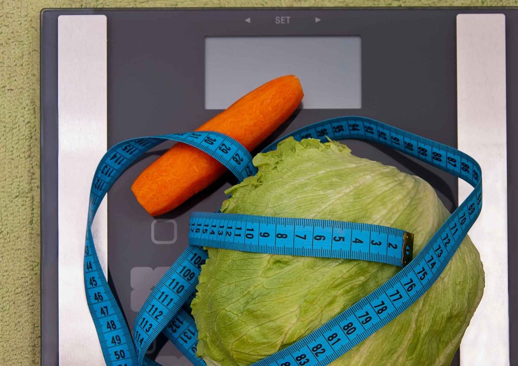What Is a Balanced Diet for Weight Loss? 13 A carrot placed on a scale next to a measuring tape, indicating its weight and length for nutritional assessment.