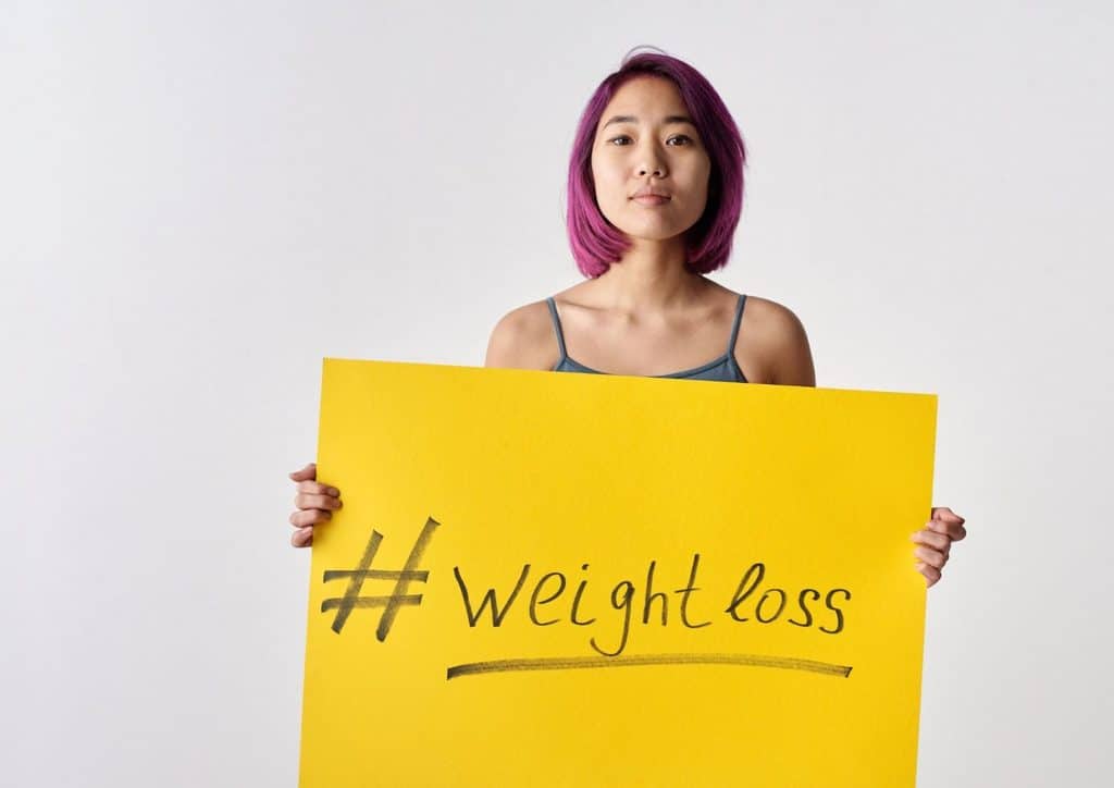 High Fibre Diet for Weight Loss: Why Fibre Keeps You Full 16 A woman stands holding a yellow sign that reads "Weight Loss," promoting health and fitness awareness.