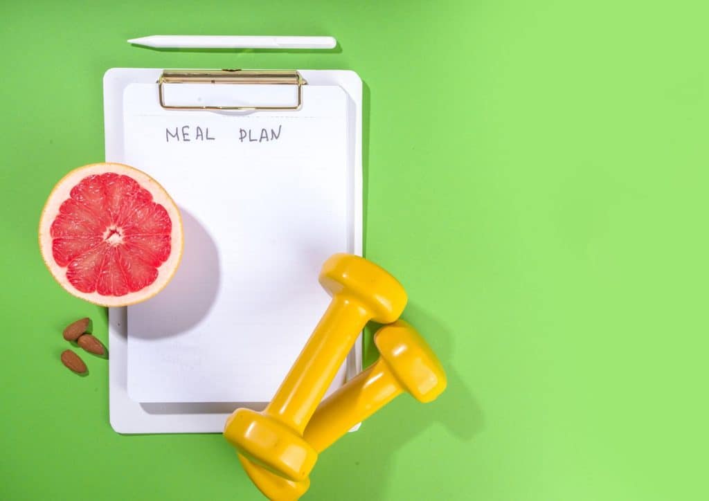Healthy Meal Plans for Weight Loss: How to Build One That Lasts 11 Clipboard displaying a meal plan, accompanied by dumbbells and a grapefruit, all on a green backdrop.