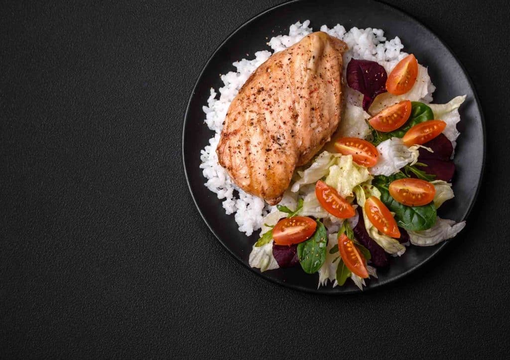 Healthy Dinners for Weight Loss: Easy Weeknight Ideas 24 A plate of grilled chicken breast accompanied by vegetables and rice, set against a black backdrop.