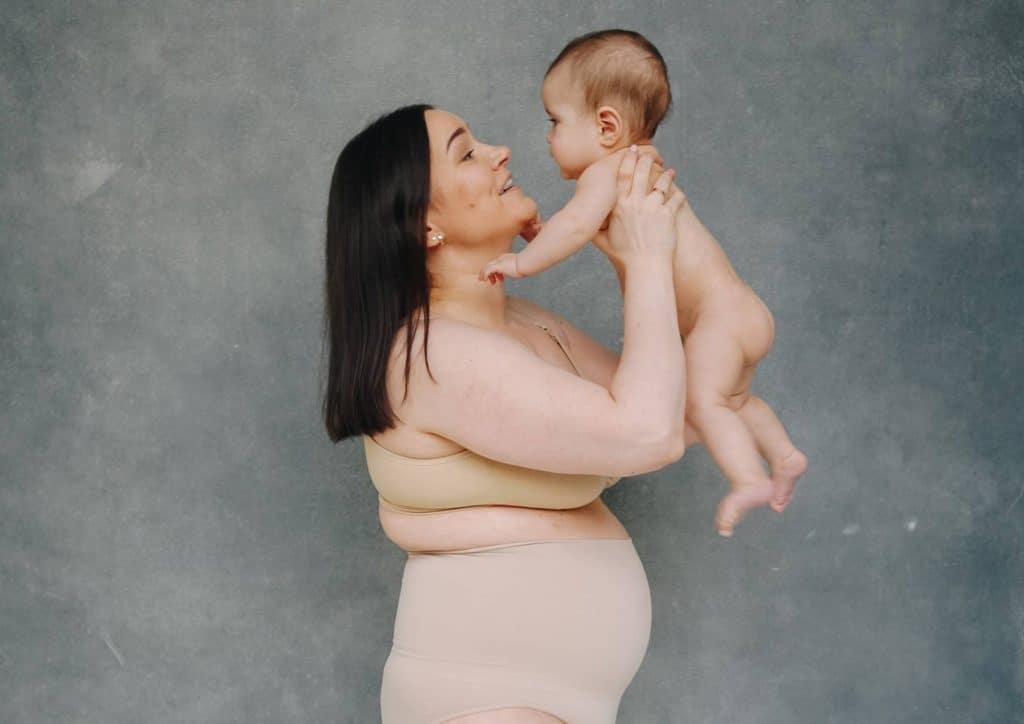 Postpartum Weight Loss: A Gentle, Realistic Guide 9 A woman in a beige bikini gently holds her baby, both smiling in a sunny outdoor setting.