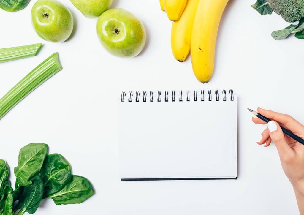 Easy Meals for Weight Loss When You're Short on Time 34 A woman's hand writes on a notepad beside a notebook filled with illustrations of various fruits and vegetables.