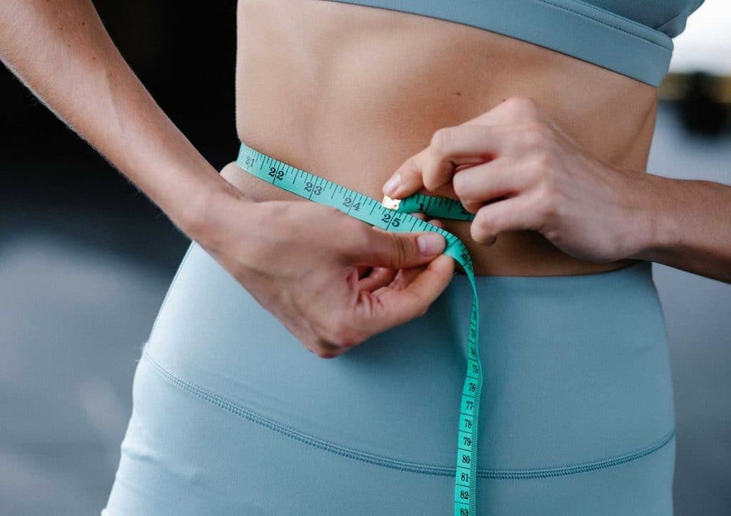High Fibre Diet for Weight Loss: Why Fibre Keeps You Full 15 A woman measuring her waist with a measuring tape, focusing on her midsection in a well-lit room.