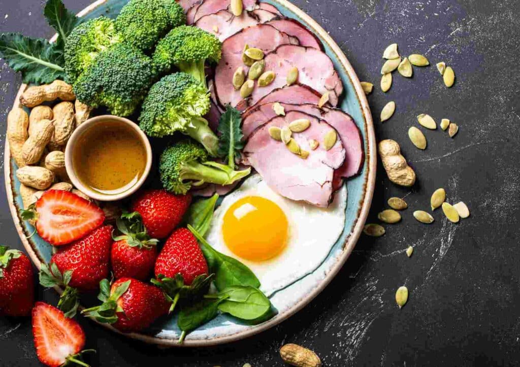 Keto vs Low Carb for Weight Loss: Which Is Better? 23 A plate featuring broccoli, strawberries, eggs, nuts, and a bowl of peanut butter, showcasing a healthy meal.