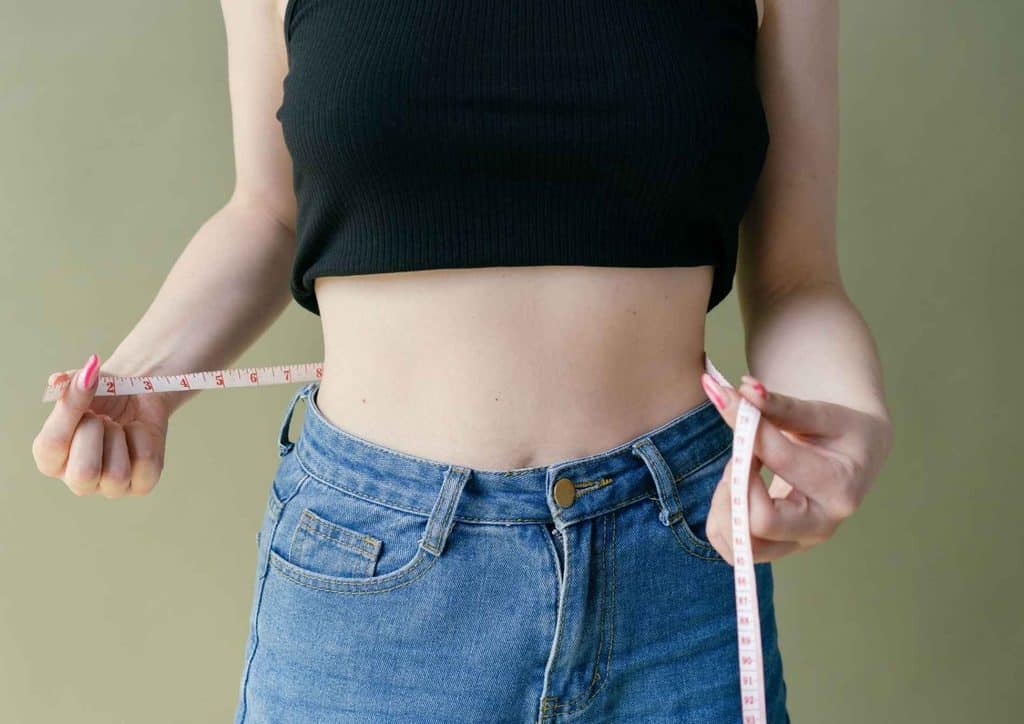 High Fibre Diet for Weight Loss: Why Fibre Keeps You Full 14 A woman measuring her waist with a tape measure, focusing on her midsection in a well-lit room.