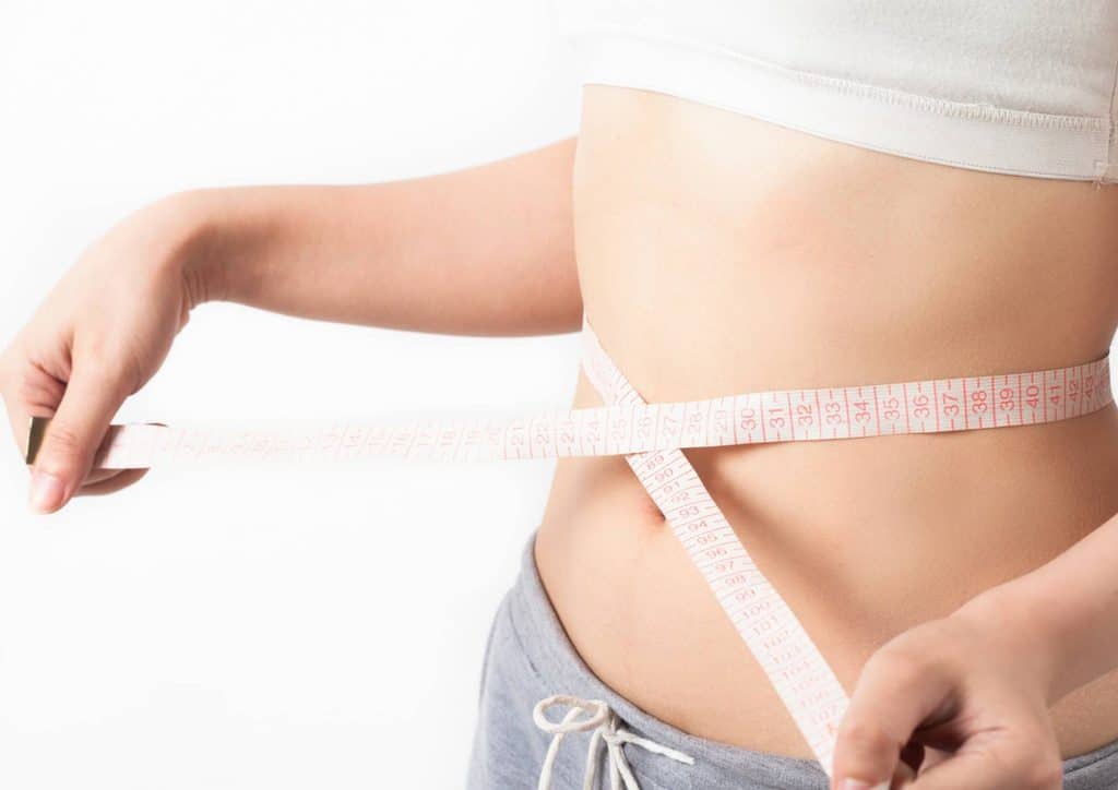 Postpartum Weight Loss: A Gentle, Realistic Guide 13 A woman measuring her waist with a tape measure, focusing on her midsection for fitness or health tracking.