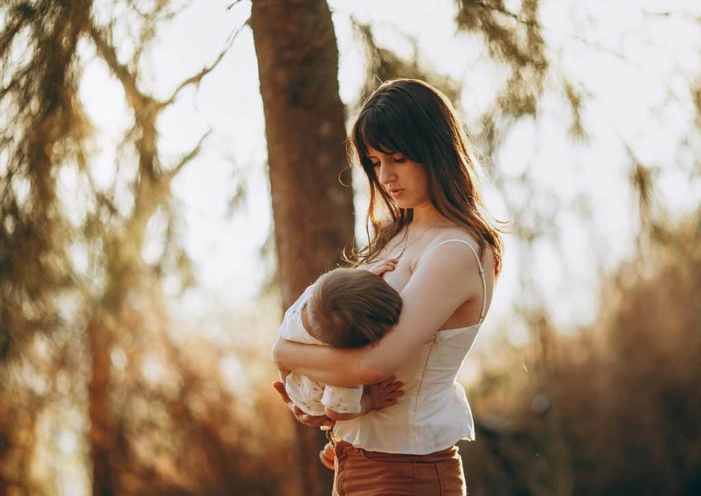 Losing Weight While Breastfeeding: What's Safe and Realistic 49 A woman cradles her baby in her arms while standing amidst a serene wooded area.