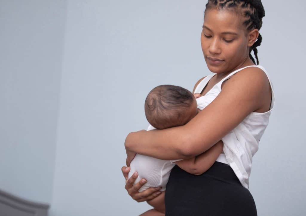 Losing Weight While Breastfeeding: What's Safe and Realistic 74 A woman stands in front of a white wall, gently holding a baby in her arms, both appearing content and serene