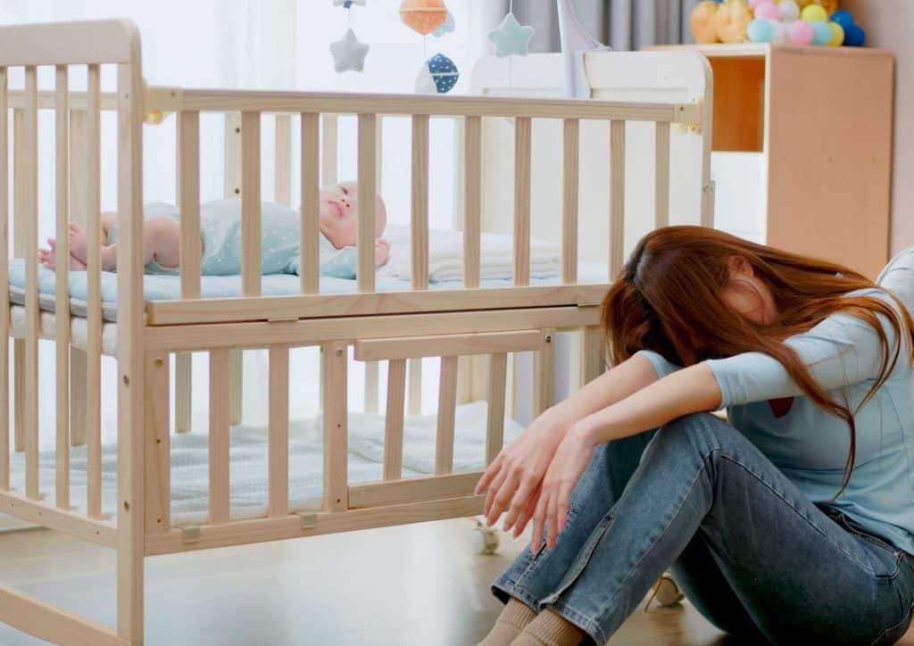 Postpartum Weight Loss: A Gentle, Realistic Guide 27 A woman sits on the floor beside a crib, looking thoughtfully at the baby inside.