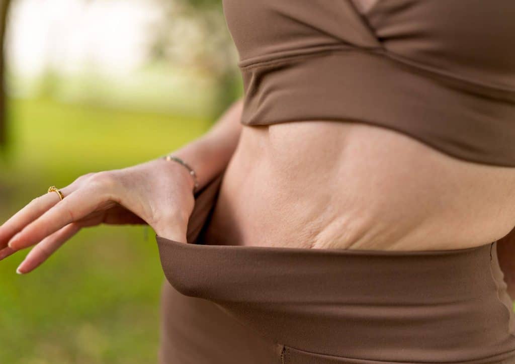 Postpartum Weight Loss: A Gentle, Realistic Guide 3 A woman in a brown top is standing and holding her stomach, appearing to be in discomfort or pain.