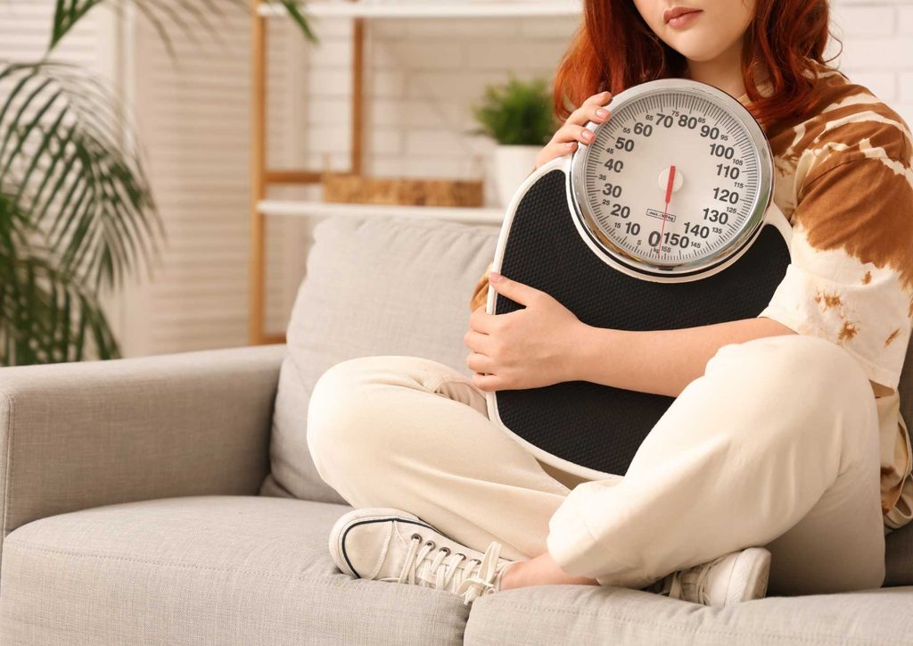 Postpartum Weight Loss: A Gentle, Realistic Guide 33 A woman sits on a couch, holding a weight scale, appearing contemplative about her health and fitness journey.