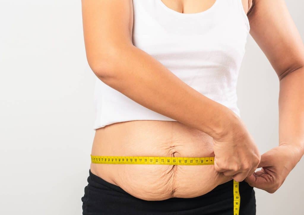 Postpartum Weight Loss: A Gentle, Realistic Guide 2 A woman measuring her waist with a tape measure, focusing on her midsection in a well-lit room.